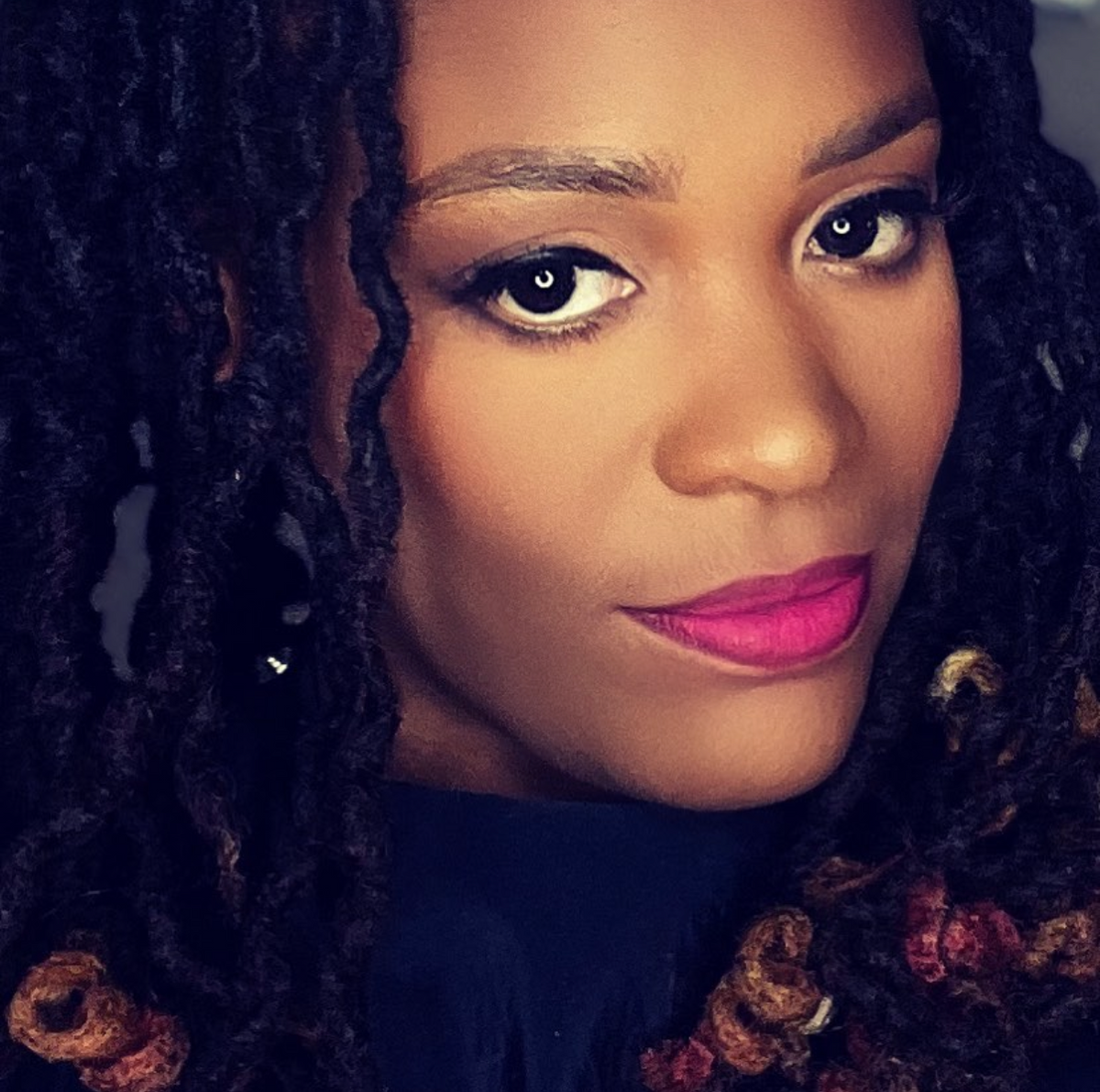 A close-up portrait of a woman with dark curly hair, wearing a dark navy top and bright pink Face Me Beauty lipstick in Shade Sistaaa