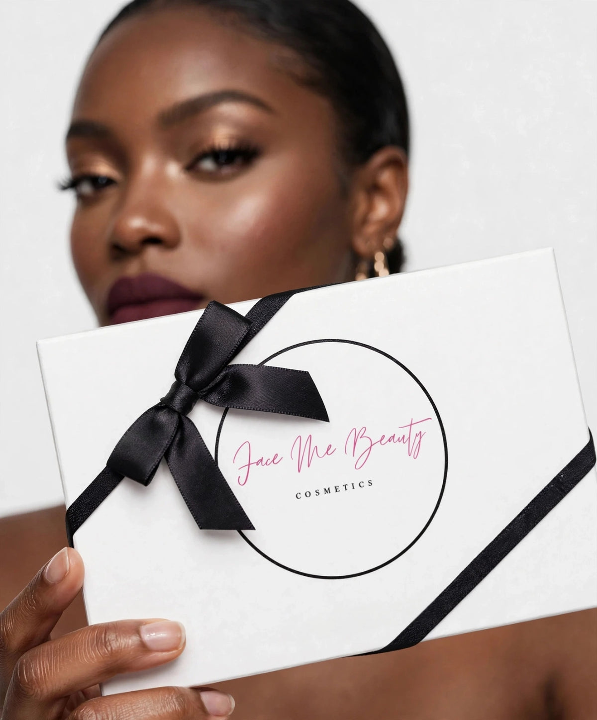 A close-up of a model holding a white cosmetic gift box with a black ribbon bow. The box features the brand name 'Face Me Beauty Cosmetics' in pink script inside a black circle.