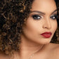 A close-up image of a woman with curly hair, featuring a natural makeup look with emphasis on the eyes, wearing a red lip, and a subtle smile.