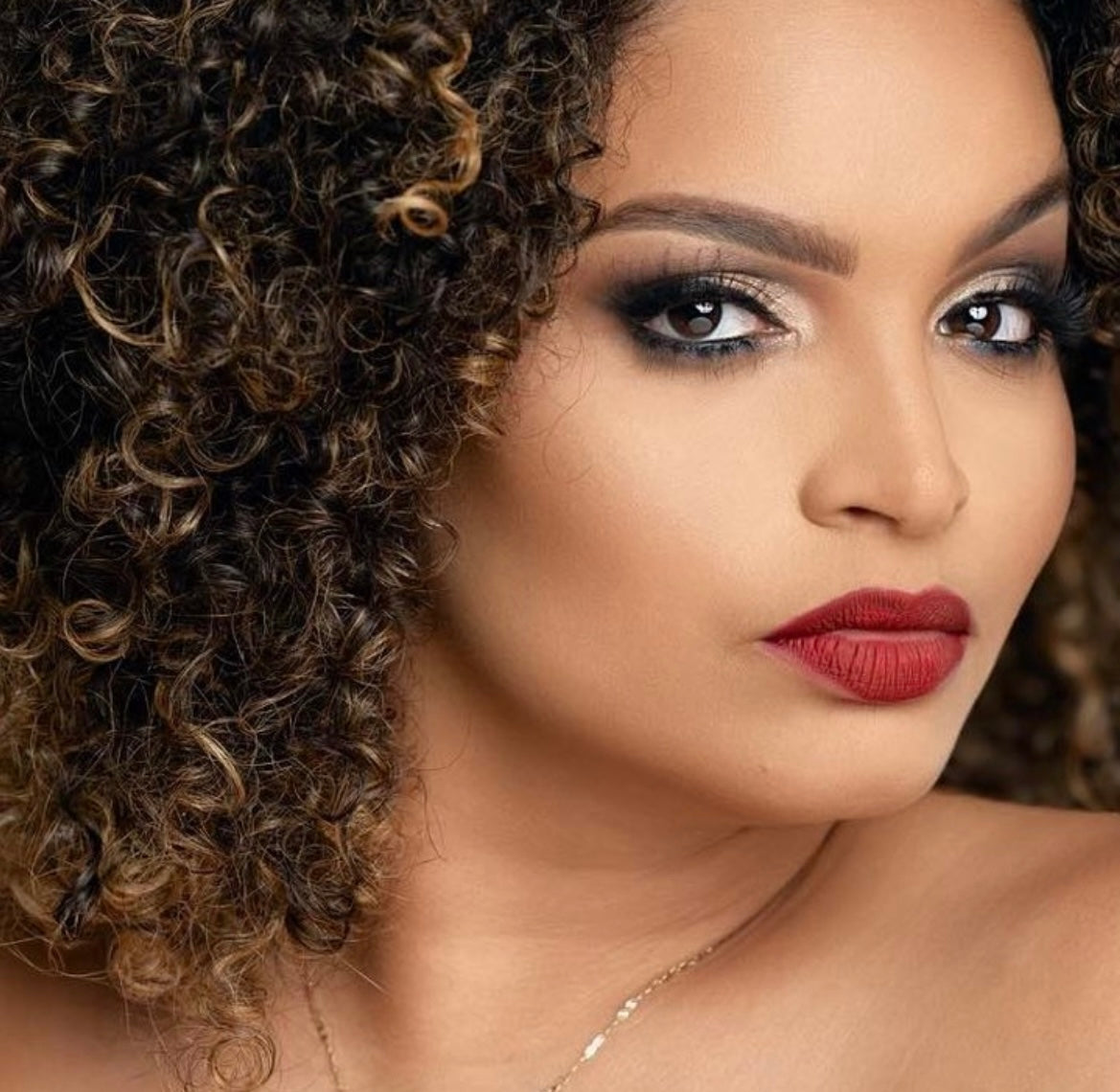 A close-up image of a woman with curly hair, featuring a natural makeup look with emphasis on the eyes, wearing a red lip, and a subtle smile.