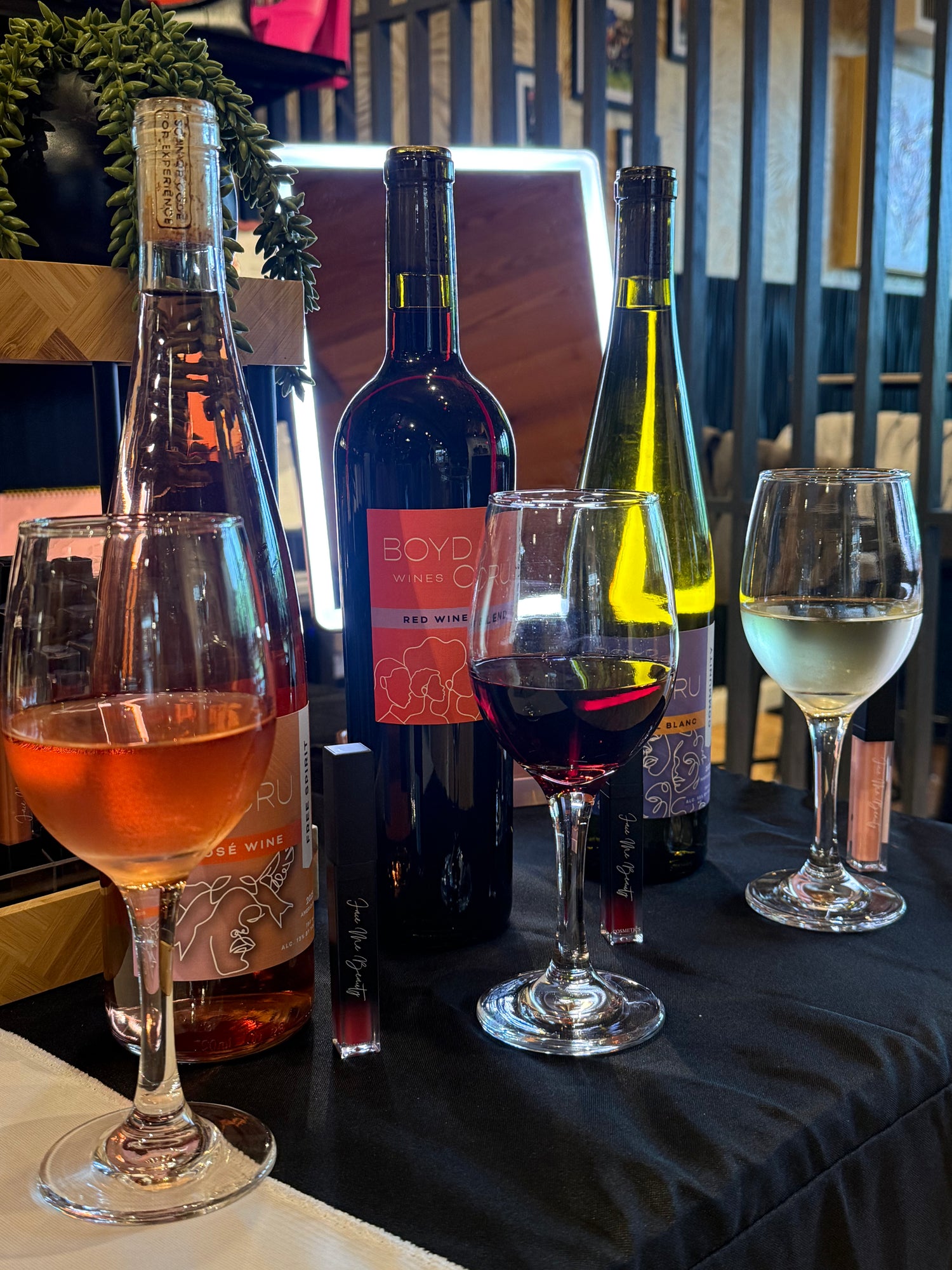 A selection of wine bottles and wine glasses arranged on a black tablecloth in an indoor setting. Visible bottles include a rosé wine bottle, a dark red wine bottle with a pink label reading 'BOYD CRU, a green wine bottle, and two wine glasses, one filled with rosé, one with red wine, and one with white wine.