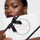 A close-up of a model holding a white cosmetic gift box with a black ribbon bow. The box features the brand name 'Face Me Beauty Cosmetics' in pink script inside a black circle.